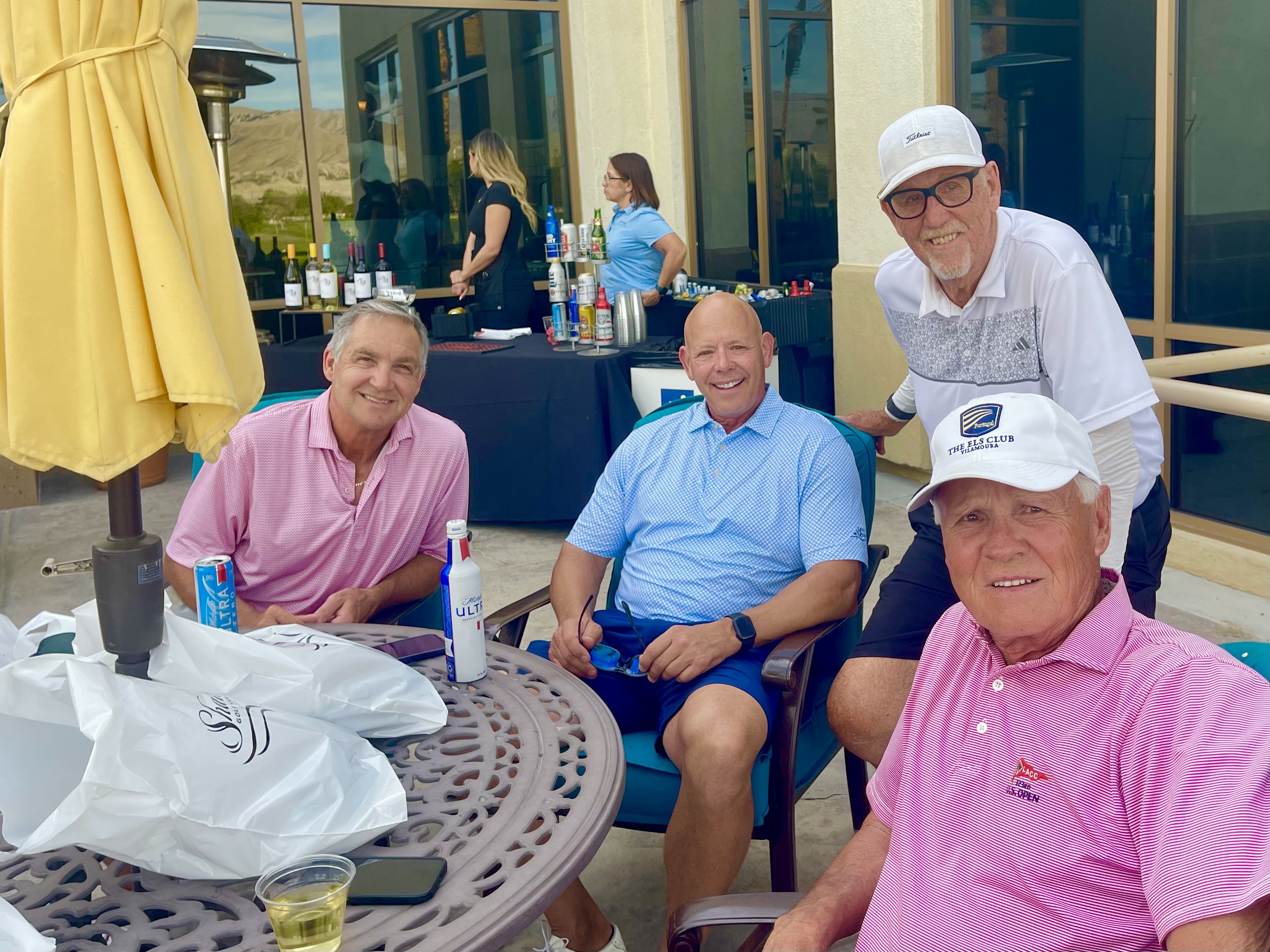 A group of people sit and relax around an outdoor table near a bar setup at a clubhouse patio.