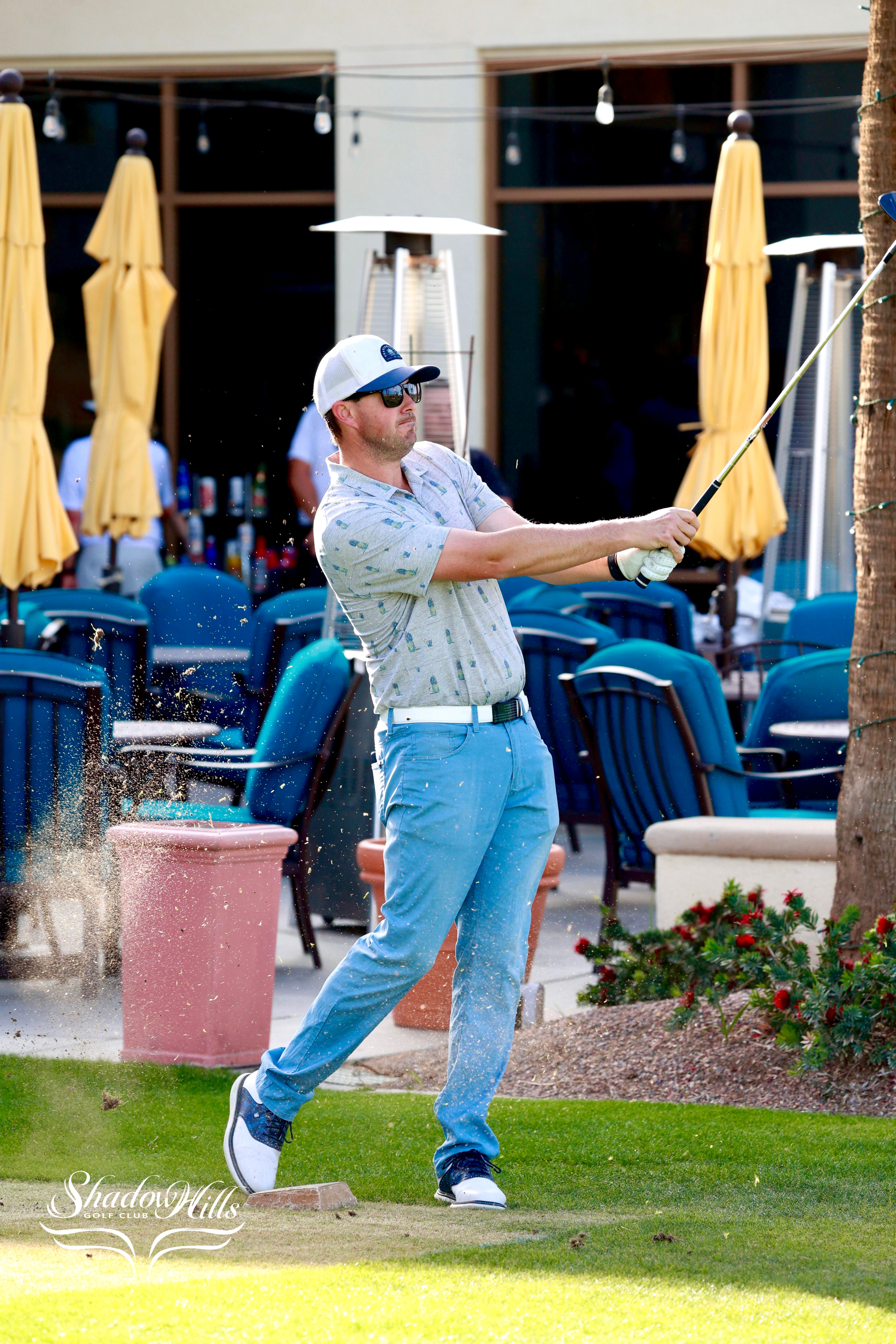 A golfer in mid‑swing hits a shot from the grass as sand sprays up behind him near an outdoor seating area.