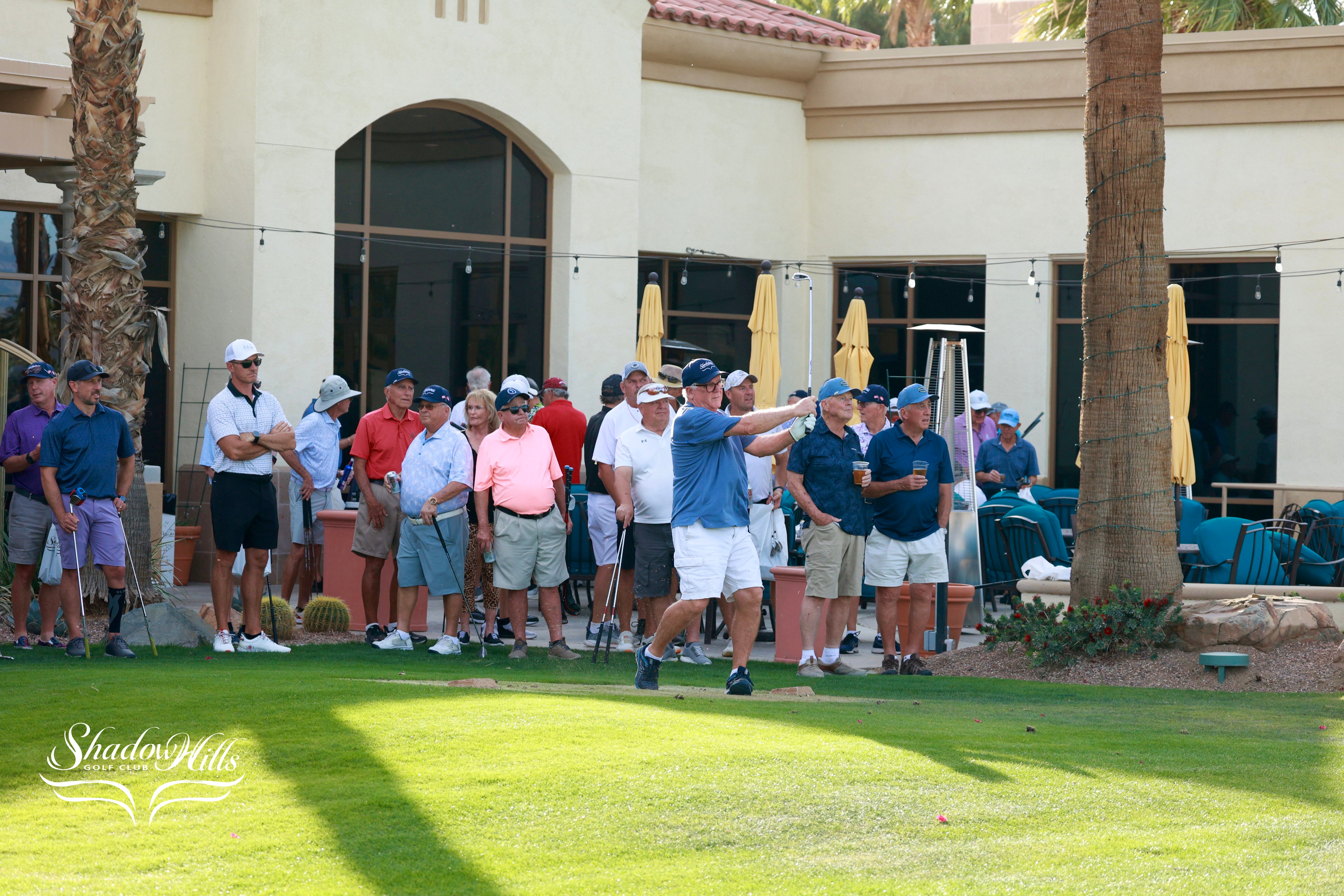 A large group of golfers stands gathered outside a clubhouse while one golfer tees off in front of them.