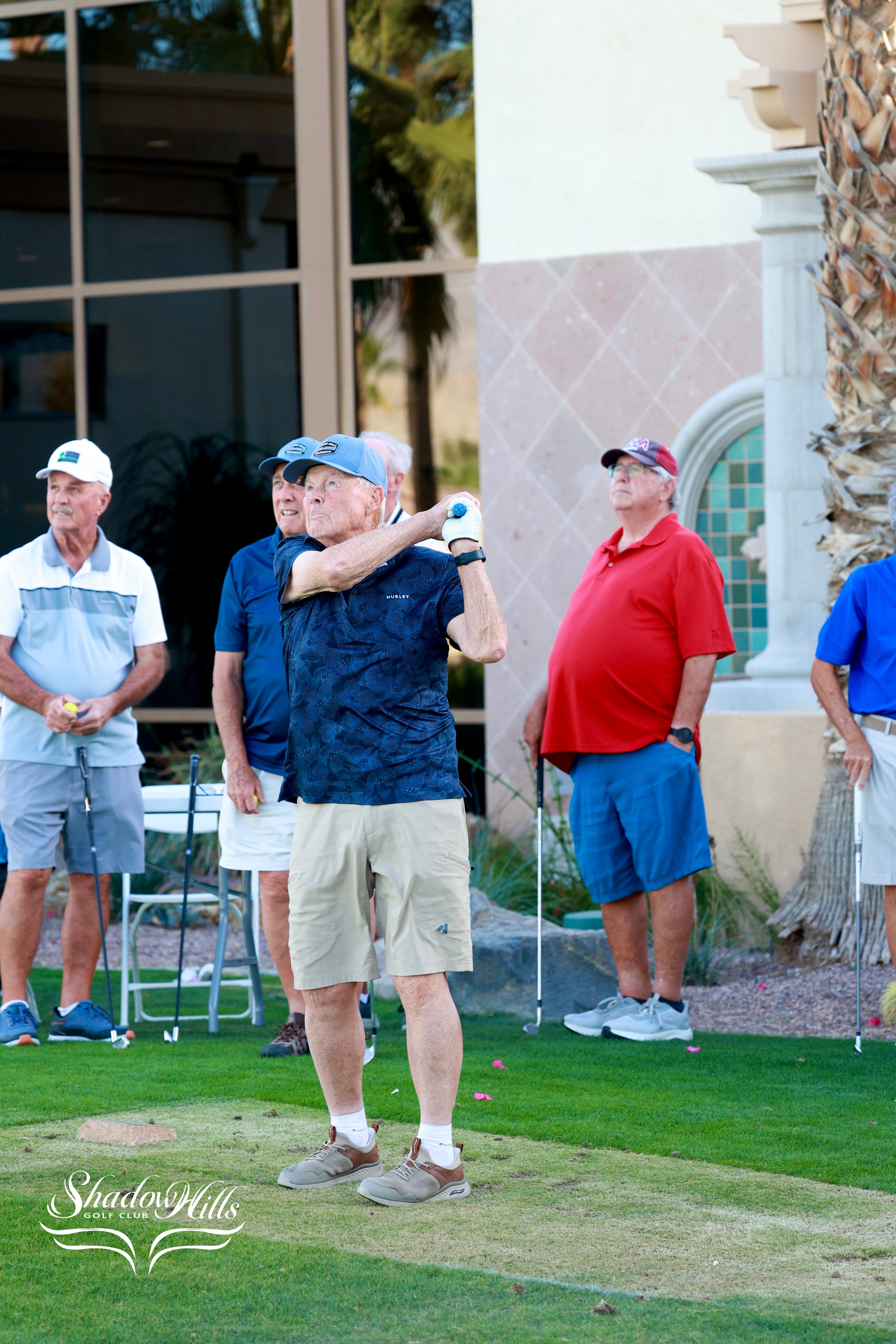 A golfer finishes his swing on the tee box as several other golfers watch from nearby.