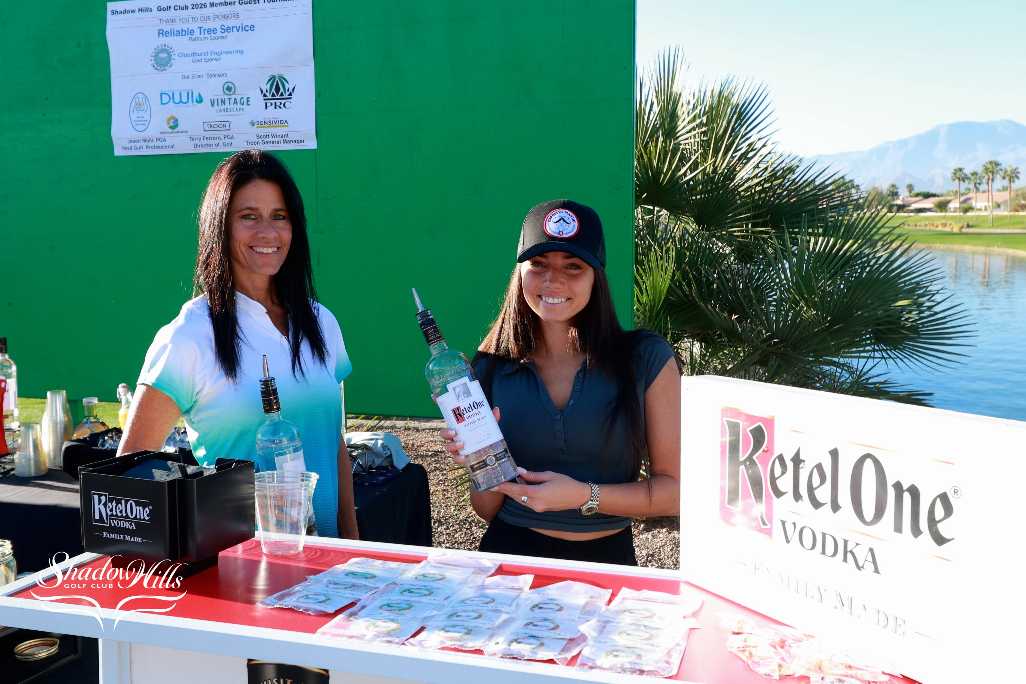 Two people stand behind a booth displaying Ketel One Vodka products at an outdoor event near a lake.