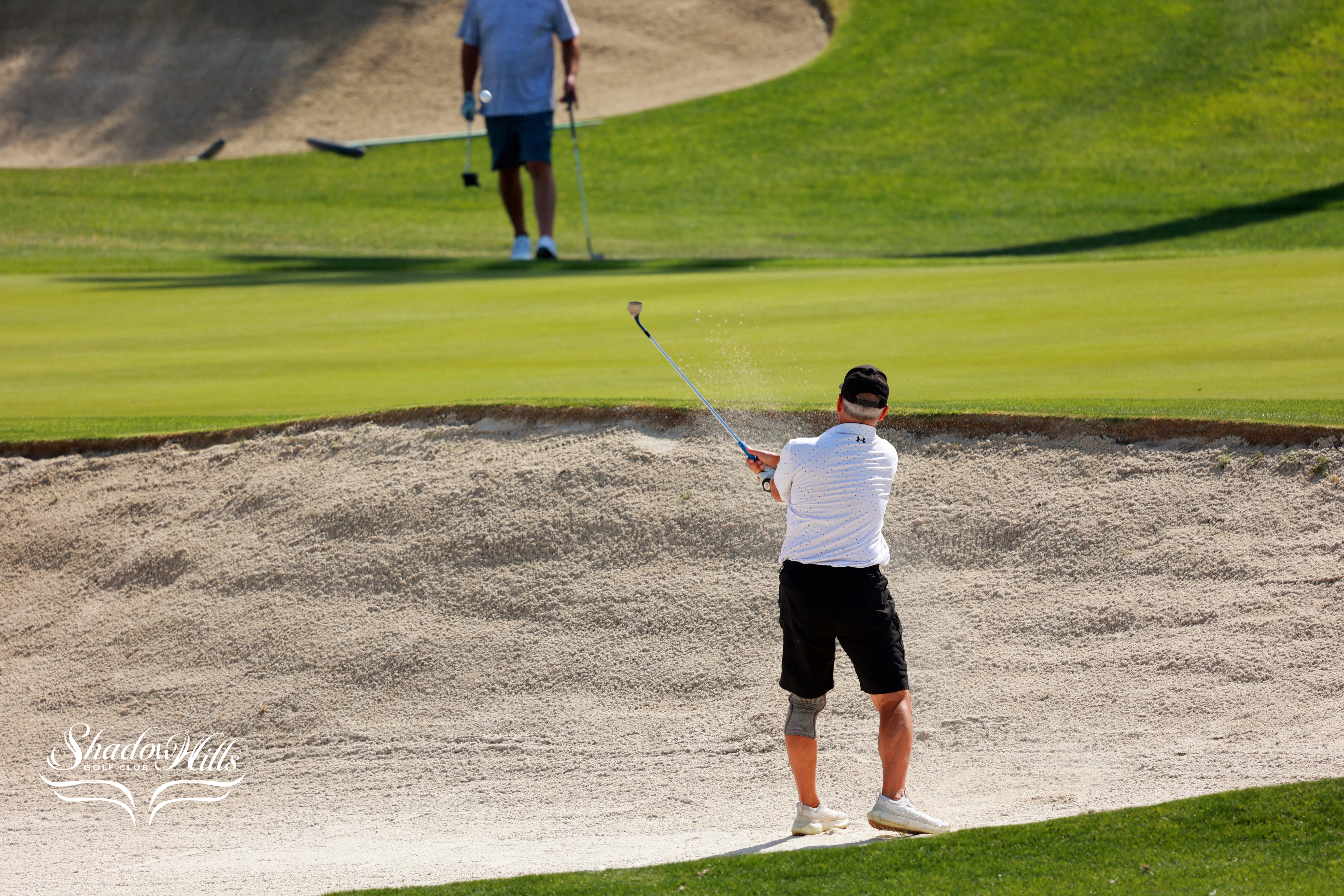 A golfer hits a shot from a sand bunker while another person stands on the green in the background.