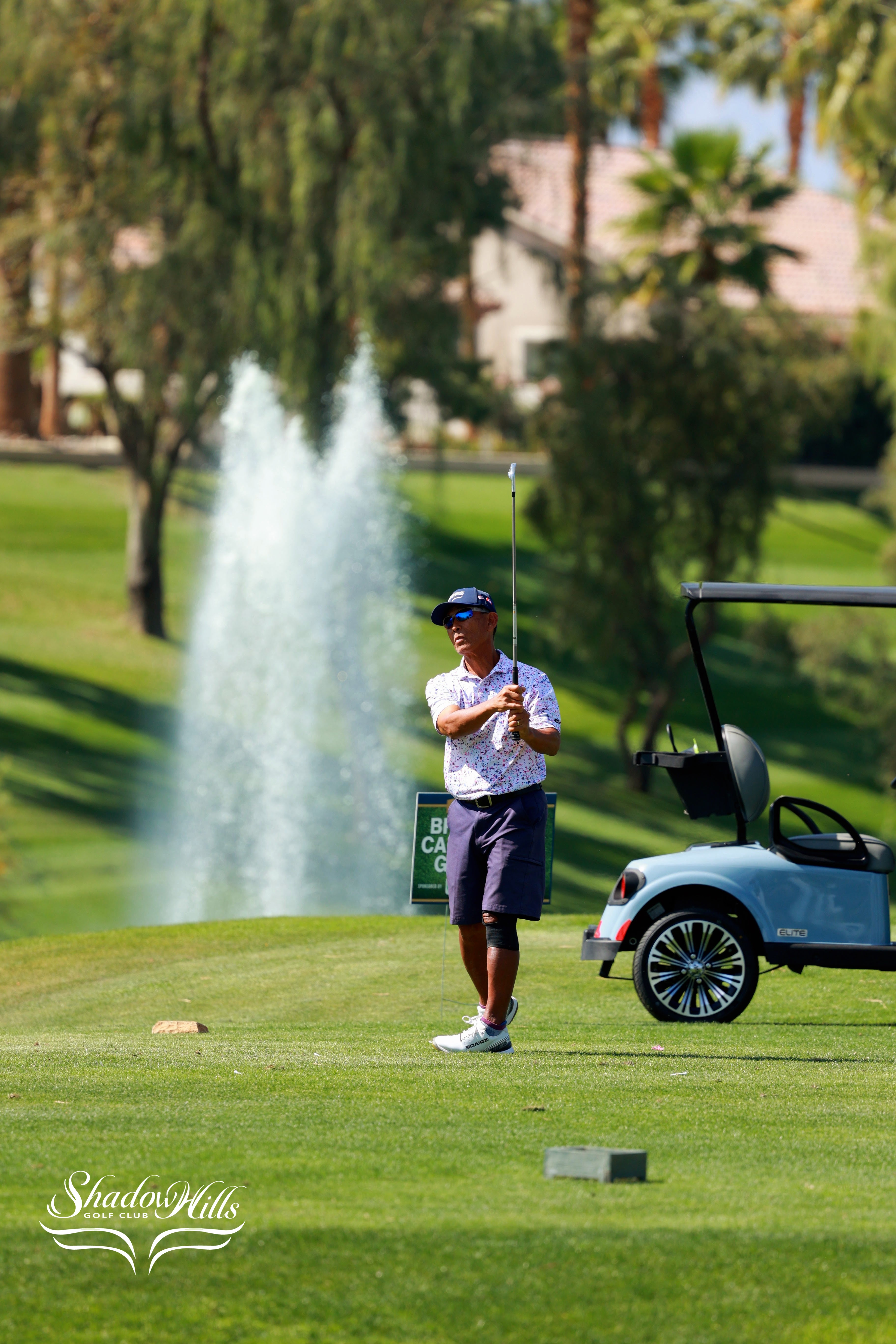 A golfer swings a club near a fountain on a lush green course with a golf cart nearby.