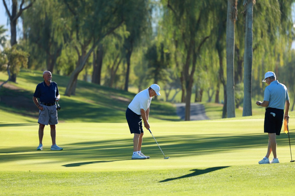 Three golfers stand on the green as one prepares to putt toward the hole.