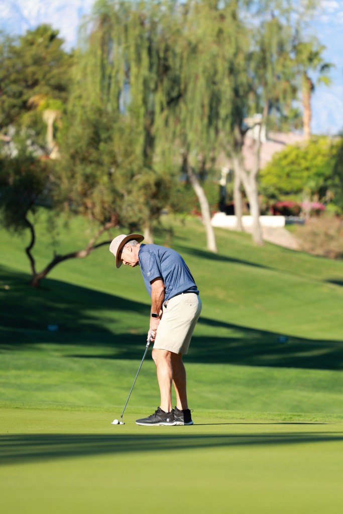 A golfer lines up a putt on a smooth green surrounded by trees and landscaped hills.