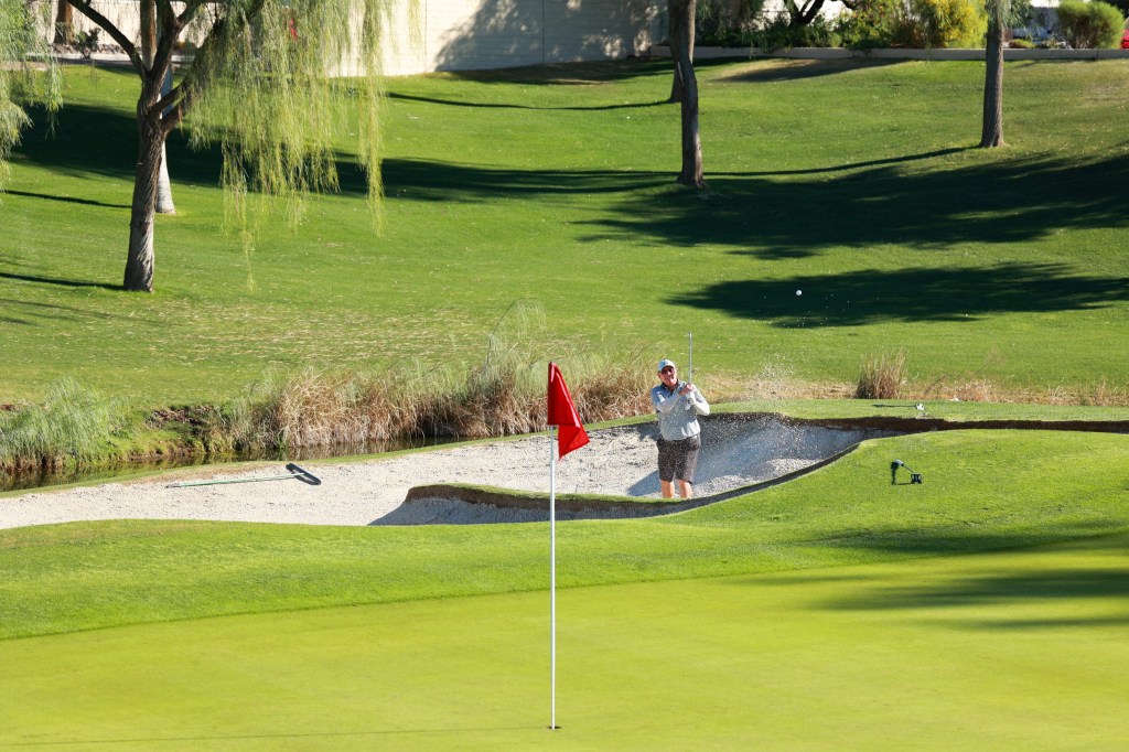 A golfer hits a shot from a sand bunker while aiming toward a green with a red flag.