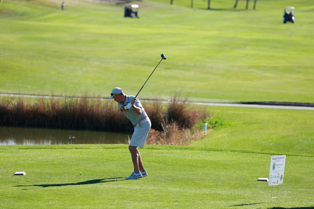 A golfer tees off near a pond with tall grass as the ball lifts from the tee.