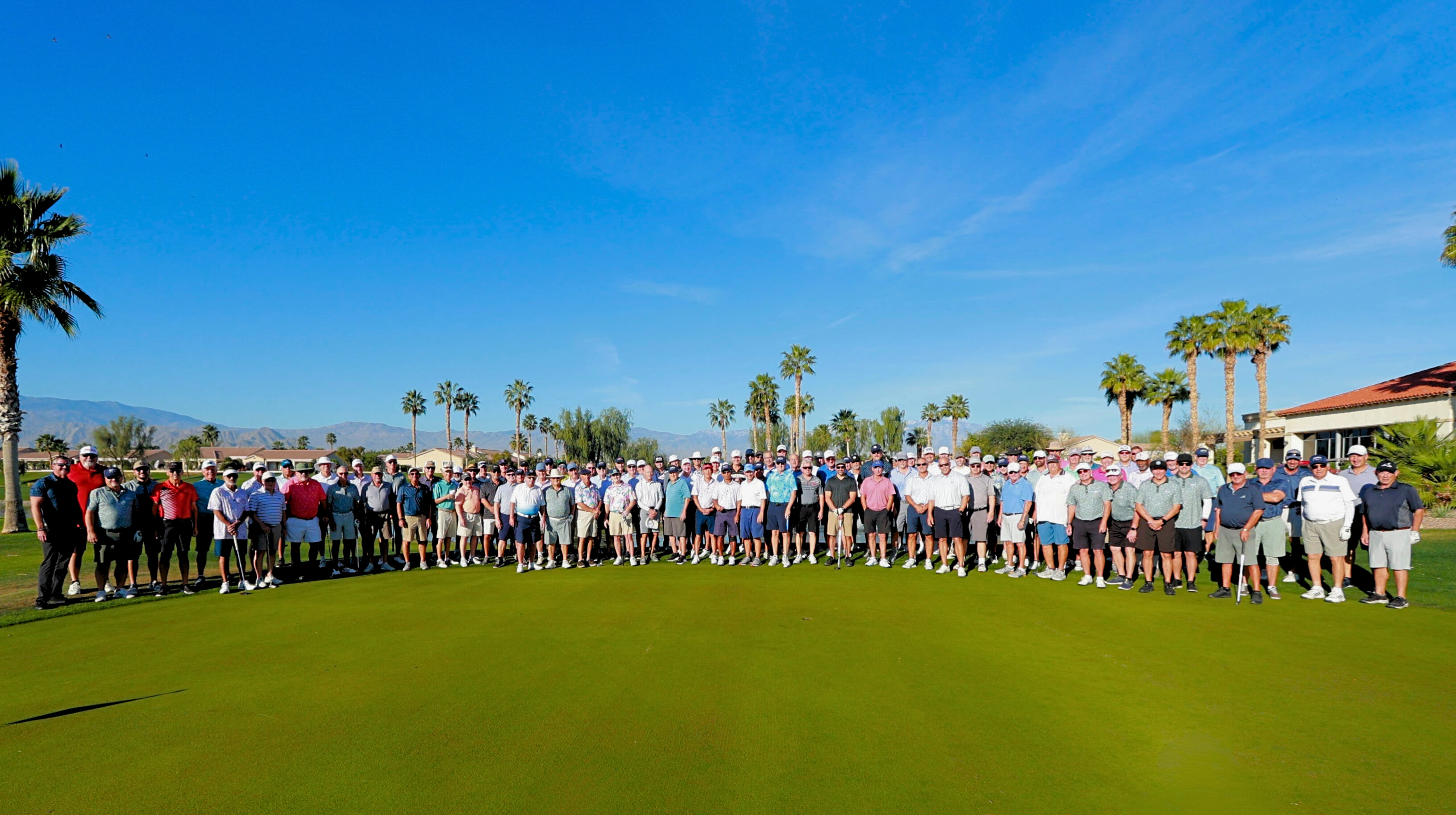 A large group of golfers stands together on a sunny golf course surrounded by palm trees.